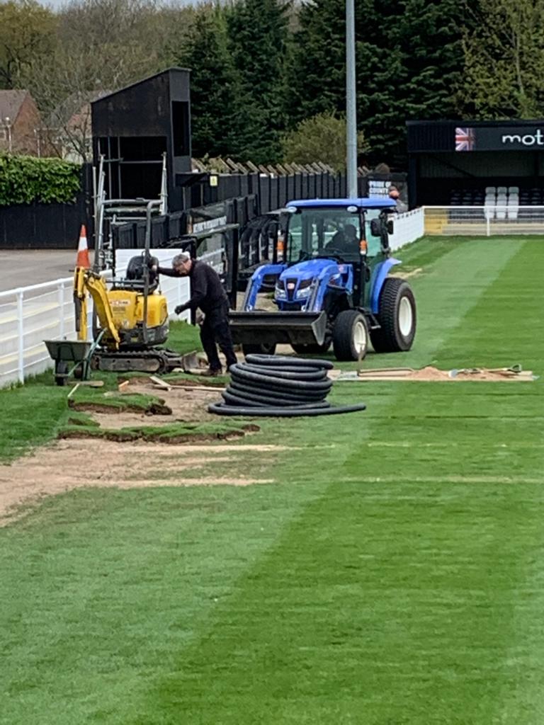 A tractor and some workmen dig shallow trenches in a pitch